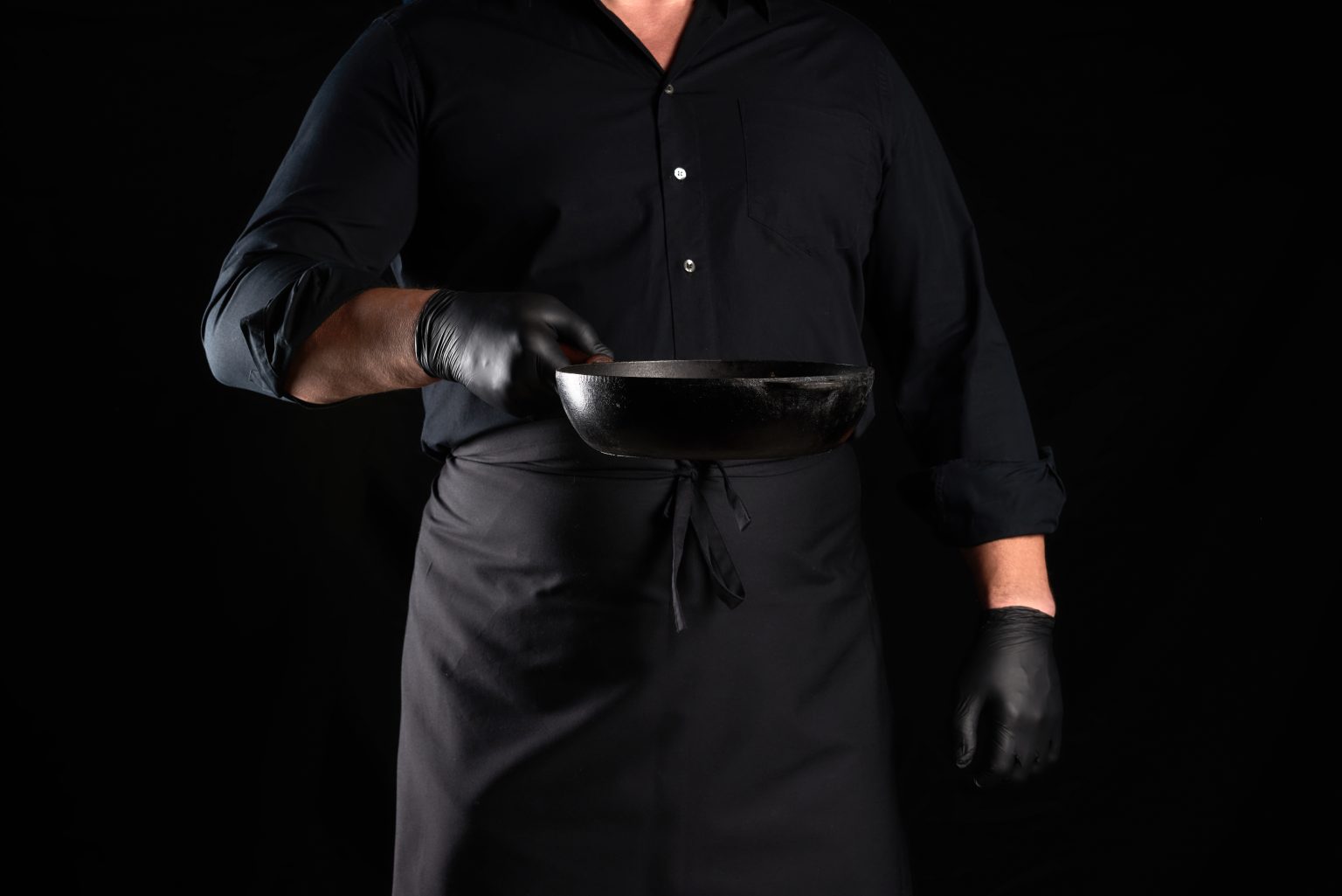 cook in black uniform and latex gloves holds an empty round vintage black cast iron pan in front of him, low key