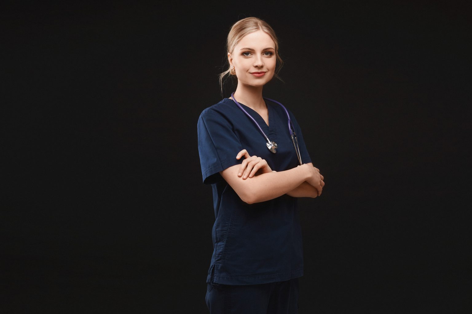 A young woman in medical uniform and with a stethoscope posing with folded hands at the dark background, isolated. Healthcare and emergency concept.